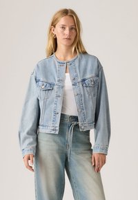 Γυναίκα που φοράει ανοιχτό μπλε τζιν μπουφάν και ξεβαμμένο τζιν ψηλόμεσο παντελόνι πάνω από ένα λευκό τοπ, στέκεται μπροστά σε απλό μπεζ φόντο.