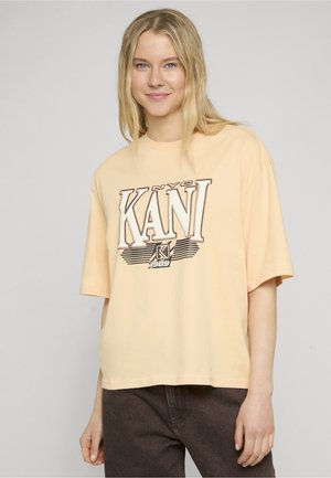 Jeune femme aux cheveux blonds porte un T-shirt oversize beige clair avec le logo "NYC KANI 1989" et un pantalon marron foncé sur un fond uni.