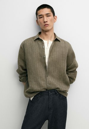 Uomo con capelli corti e scuri indossa una camicia verde a coste con bottoni sopra una maglietta henley bianca e jeans scuri, in piedi davanti a uno sfondo chiaro uniforme.
