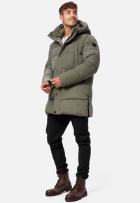 Olive green puffer jacket with a hood, featuring a quilted design, snap buttons, and a patch on the sleeve, paired with black pants and brown boots.