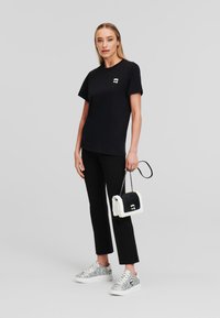 T-shirt noir à manches courtes avec un petit logo blanc, pantalon noir ajusté, baskets argentées et un sac noir et blanc.