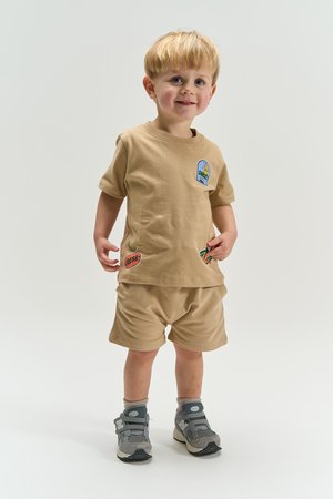Väikelaps naeratab, kannab beežikaid lühikesi pükse ja t-särki värvikate plaastritega, hallid vabaajajalatsid, seisab ühevärvilise heleda tausta ees.