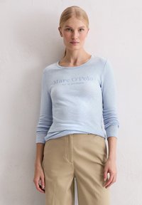 Lichtblauw T-shirt met lange mouwen en ronde halslijn, voorzien van de opdruk "Marc O'Polo EST. IN STOCKHOLM"; gecombineerd met beige broeken.