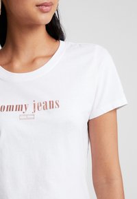 Žena na sobě má bílé tričko s krátkým rukávem a kulatým výstřihem, na hrudi je v světle hnědé barvě natištěné "Tommy jeans".