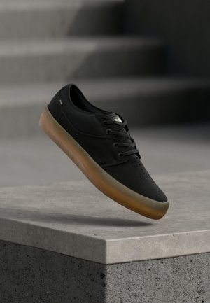 Black casual lace-up shoe with brown sole floating above a concrete ledge with blurred stairs in the background.