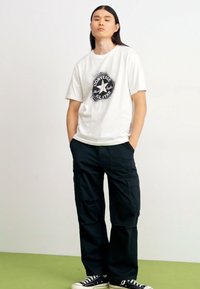 Camiseta blanca de manga corta con un logo negro de Converse All Star, combinada con pantalones cargo negros y zapatillas en blanco y negro.