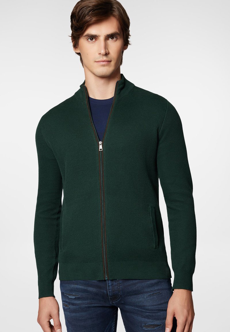 Zielony sweter zapinany na zamek z teksturowanej dzianiny, z kołnierzem typu stójka, bocznymi kieszeniami i kontrastowym detalem zamka. Nosi się go na granatowej koszuli.