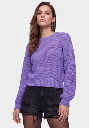 Jonge vrouw met lang haar draagt een paarse gebreide trui, zwarte geripte denimshorts en stervormige hangende oorbellen tegen een effen achtergrond.
