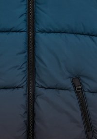 Tamsiai mėlynas pūkinis švarkas su centriniu užtrauktuku, turinčiu tekstūruotą traukimo rankenėlę ir užtrauktuku uždaroma kišene dešinėje pusėje.