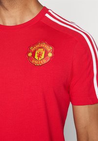 Piros pamut póló kerek nyakkal, a vállakon fehér csíkokkal és hímzett Manchester United logóval a mellkasán.