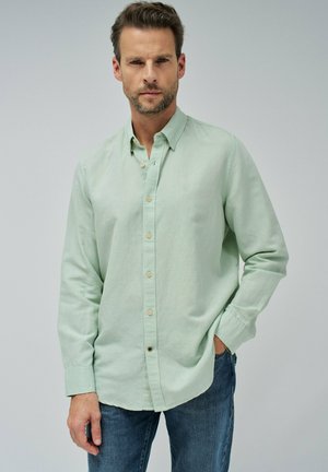 Hombre con una camisa de botones de manga larga verde claro y jeans azules, de pie con una mano en el bolsillo frente a un fondo liso.