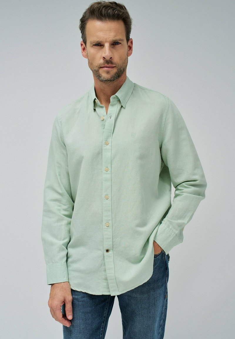 Homme portant une chemise à manches longues vert clair et un jean bleu, se tenant avec une main dans la poche devant un fond uni.