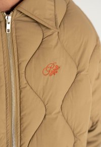 Beige puffatakki, jossa on tikattu muotoilu, varustettuna vetskarilla ja hupulla. Vetskarin lähellä on oranssi brodeerattu logo.