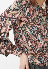 Blouse à manches longues en tissu sombre, ornée d'un motif floral paisley vibrant avec des accents rose, teal et crème. Texture douce, manches froncées.