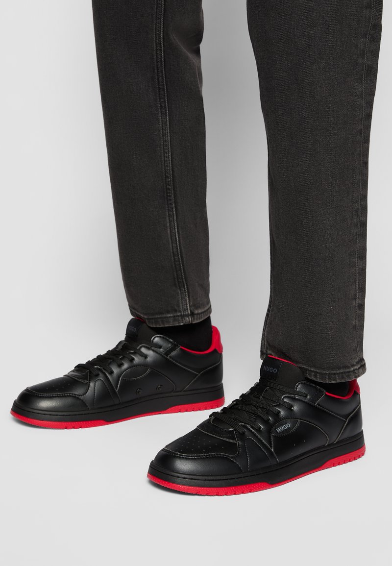 Baskets noires avec des accents rouges, tige en cuir, bout rond, col rembourré, semelle extérieure texturée, fermeture à lacets et détail de logo sur le côté.
