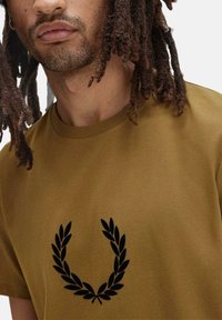 T-shirt in cotone marrone con logo a corona d'alloro nero sul petto. Il design è semplice, con collo rotondo e vestibilità rilassata.