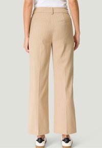 Pantalon beige à larges rayures avec une texture lisse, doté de poches arrière et d'une taille mi-haute, associé à des baskets blanches.