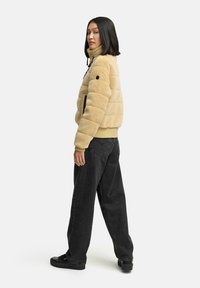 Beige Kunstfelljacke mit hohem Kragen, gestepptem Design und zwei seitlichen Taschen, kombiniert mit lässigen schwarzen Jeans und schwarzen Sneakers.