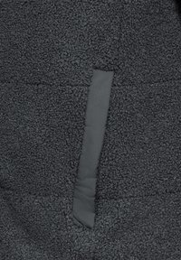Giacca in pile nera con una superficie testurizzata e morbida. Presenta un pannello in tessuto grigio a contrasto su un lato, che arricchisce il dettaglio del design.