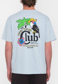 Camiseta de color azul claro con un gráfico colorido que presenta un tucán, hojas de palma y un diseño floral, incluyendo texto, en la parte de atrás.