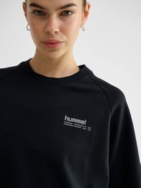 Junge Frau mit goldenen Creolen, die einen schwarzen Hummel-Sweatshirt mit Logo und kleinem Text auf der linken Brust vor hellem Hintergrund trägt.