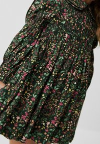 Abito floreale in verde scuro con texture arricciata, caratterizzato da fiori rosa, gialli e bianchi; ha un colletto rotondo e maniche a sbuffo.