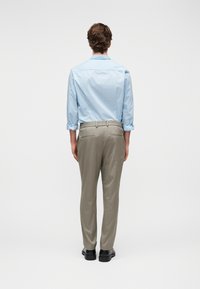 Άνδρας που φοράει ανοιχτό μπλε πουκάμισο με μακριά μανίκια και μπεζ παντελόνι στέκεται με την πλάτη προς τα εμπρός σε απλό λευκό φόντο.