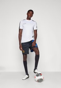 Chemise de sport à manches courtes blanche avec des accents noirs, associée à un short noir comportant un design et un logo bleus, des chaussettes noires et des crampons blancs.