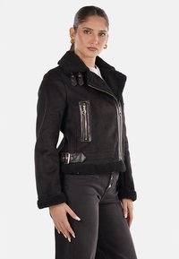 Giacca nera in scamosciato sintetico con colletto in shearling, zip e tasche decorative, e vita aderente. Presenta una chiusura decorativa con zip e cintura.