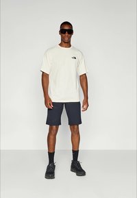 Light beige t-shirt with a black logo, paired with dark navy shorts and black trail shoes. Model wears sunglasses.