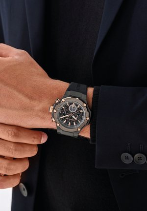 Montre chronographe noire avec des accents en or rose, bracelet en caoutchouc et un cadran noir texturé affichant divers cadrans et repères.
