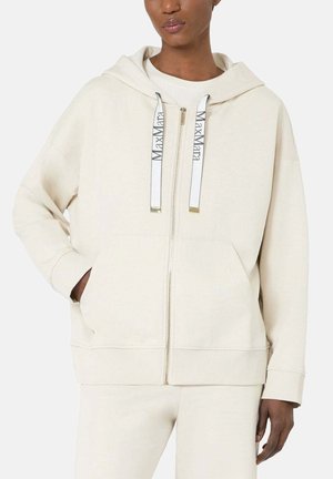 Persona che indossa una felpa crema con zip, grandi tasche frontali e coulisse bianche stampate con il testo "Max Mara".