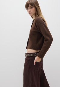Cárdigan corto marrón de punto suave con puños y dobladillo acanalados, combinado con pantalones marrones oscuros que tienen un cinturón con ojales metálicos.