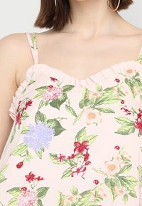 Top sin mangas floral con un fondo rosa suave, hojas verdes y flores multicolores. Detalles de escote con volantes y tirantes finos.