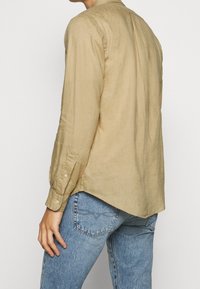 Osoba v béžové lněné košili s dlouhým rukávem, kterou má vpředu vytaženou, nad světlými modrými džíny, zobrazená zezadu a z boku.