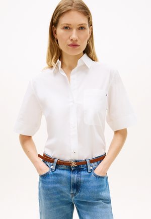 Žena s rukami v vreckách, oblečená v bielej krátkorukáčovej košeli zapínanej na gombíky, modrých džínsoch, hnedom opasku a zlatých kruhových náušniciach.