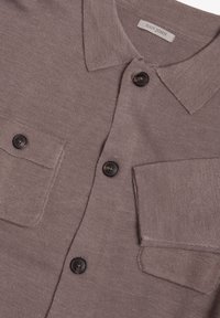 Beige strikket cardigan med krave, to brystlommer og fire sorte knapper. Stoffet har en glat tekstur og subtil ribbning.