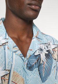Camicia a maniche corte in tessuto azzurro chiaro con stampa floreale tropicale, bottoni e colletto rilassato. Texture con uno sfondo a motivi.