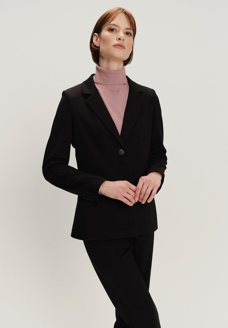 Zwarte op maat gemaakte blazer met een enkele knoopsluiting, gedragen over een lichtroze coltrui. Gladde stof met gestructureerde schouders en aansluitende mouwen.