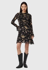 Vestido negro con estampado floral, mangas largas, corpiño fruncido, falda con volantes y textura ligera. Combinado con botas de vaquero negras para contraste.