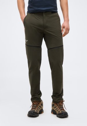 Homme portant un pantalon de randonnée convertible vert foncé et des chaussures de randonnée marron, debout les mains détendues le long du corps.