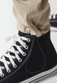 Zapatilla alta de lona negra con cordones y costuras blancas, combinada con pantalones beige ajustados. Cuenta con una suela de goma y ojales de metal.