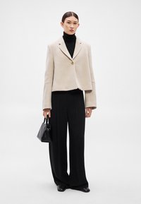 Μπεζ cropped σακάκι με δομημένους ώμους, κλείσιμο με ένα κουμπί· συνδυασμένο με μαύρη ζιβάγκο και φαρδιές μαύρες παντελόνες.