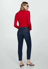 Maglietta rossa a maniche lunghe, aderente in vita, abbinata a jeans scuri a vita alta. La modella indossa sandali neri e occhiali da sole. Sfondo pulito.