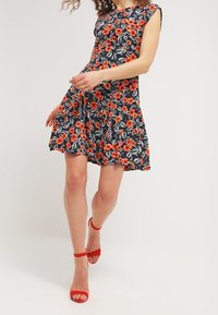 Květinové šaty s tmavě modrým pozadím, které zdobí červené květy a zelené listy. Krátké rukávy a rozšířená sukně. Doplněné červenými sandály na podpatku.