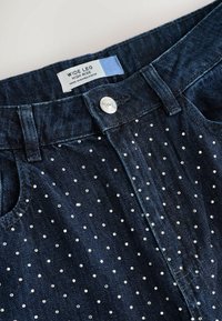 Plati kojų, aukšto liemens džinsai tamsiai mėlynos spalvos, su taškinio rašto metaliniais kalnų akmenimis, užbaigti metaliniu mygtuko uždarymu.