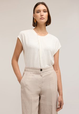 Femme portant un chemisier blanc sans manches et un pantalon beige taille haute avec un seul bouton noir, debout avec une main dans la poche.