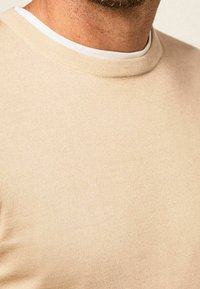 Pull en coton beige avec une texture côtelée et un col rond. Le col intérieur présente un accent blanc autour de la zone du cou.