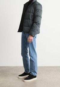 Giacca imbottita verde scuro con design trapuntato, chiusura a zip e una toppa sulla manica. Abbinata a jeans azzurri chiari e scarpe nere.