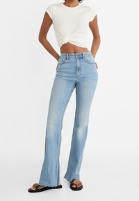 Lichtblauwe uitlopende jeans met een hoge taille, rafelige zoom en subtiele vervaging, gecombineerd met een witte geknoopte crop top en zwarte sandalen.